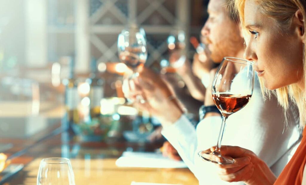 People seated at a bar, holding and tasting wine glasses, with bottles and glasses in the background.