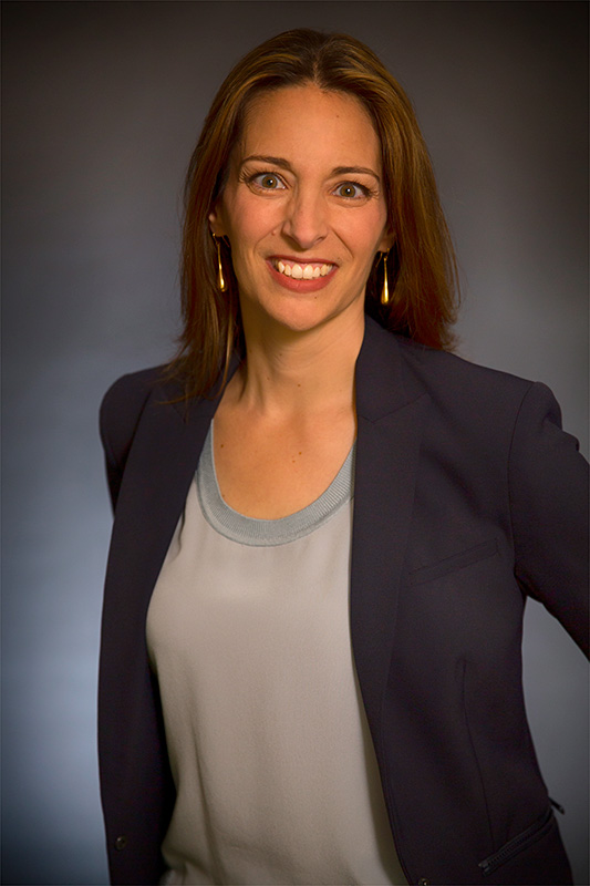 A woman with long brown hair wearing a navy blazer and light blue top smiles in a professional headshot.