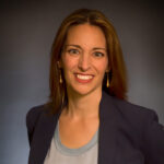 A woman with shoulder-length brown hair, wearing a dark blazer and light-colored shirt, smiles in front of a plain dark background.