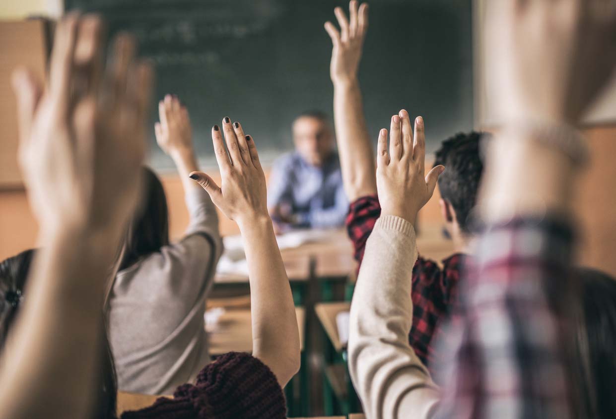 Students in a classroom raise their hands, presumably to answer or ask questions. A teacher sits at the front desk, partially blurred in the background.