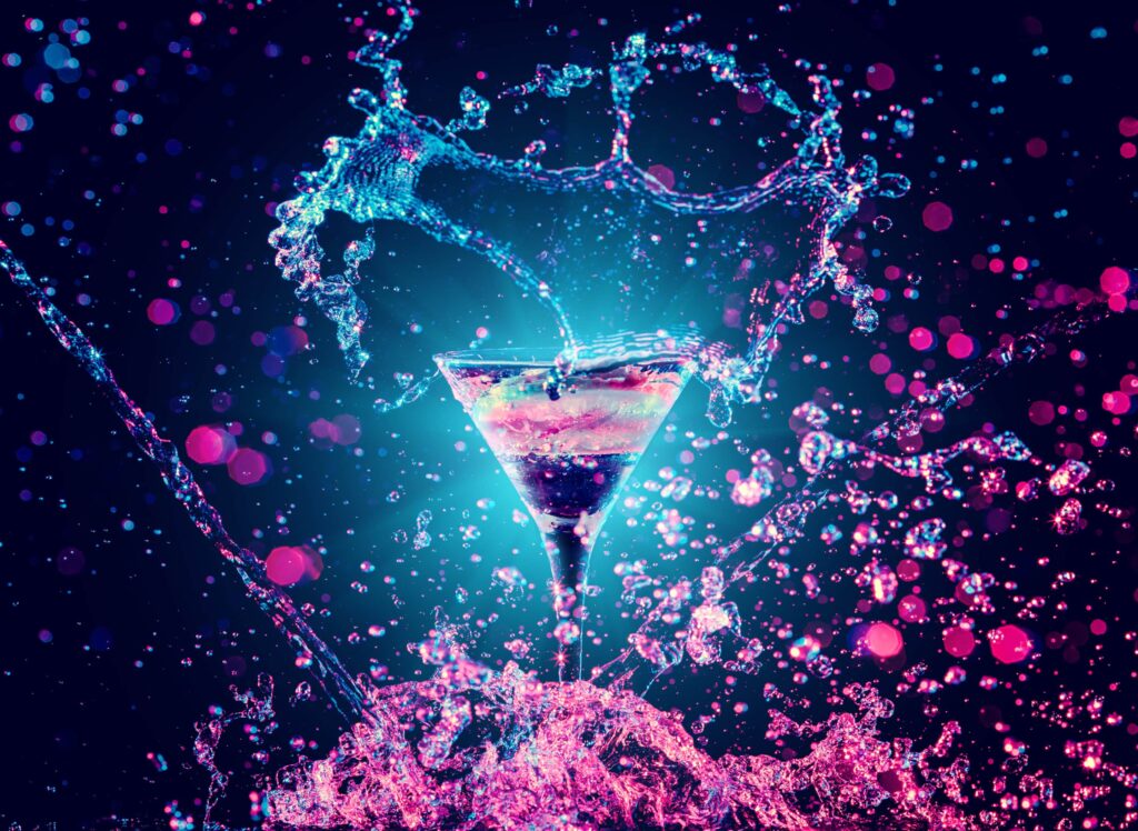 A cocktail glass is surrounded by splashes of liquid and vibrant lights with pink and blue hues against a dark background.