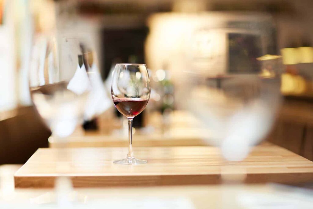 A glass of red wine sits on a wooden table, surrounded by empty glasses, in a blurred cafe or restaurant setting.