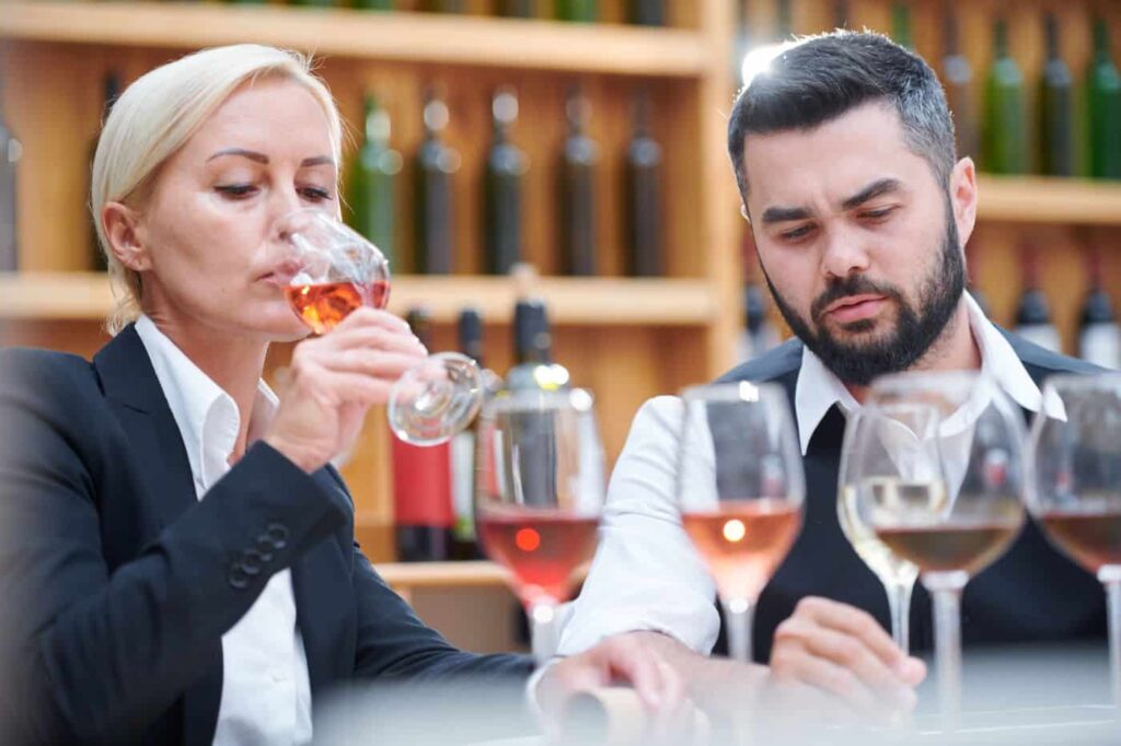 Two people are tasting wine at a wine bar, surrounded by glasses of various types of wine. Shelves filled with wine bottles are visible in the background.