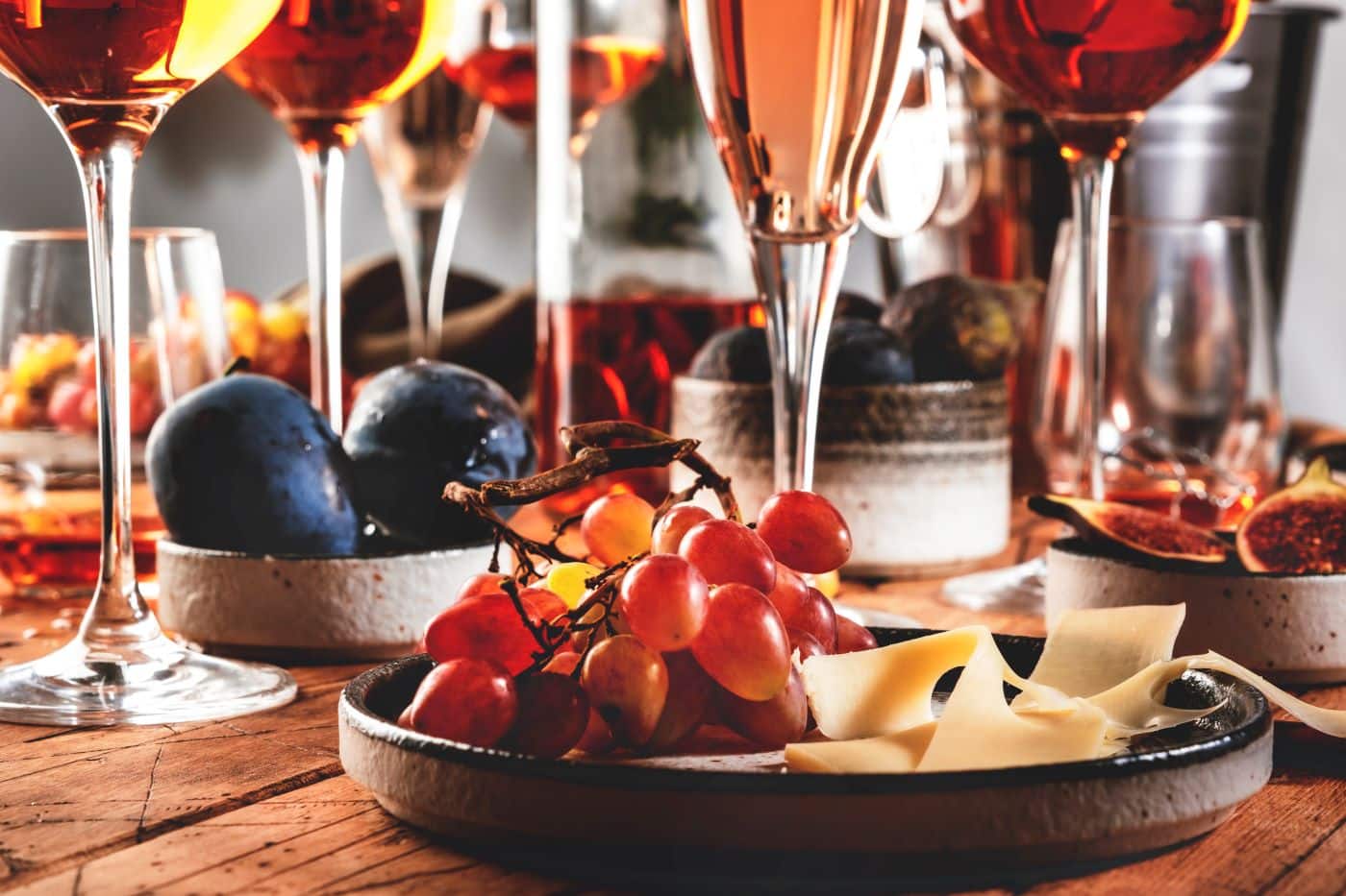 A rustic table setting with glasses of wine, a plate of grapes and cheese, bowls of figs, and plums.