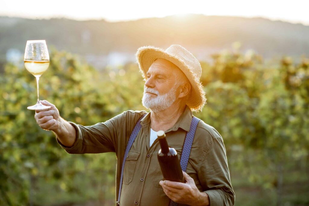 An elderly man in a straw hat holds up a glass of white wine, examining it closely, while standing in a vineyard at sunset and holding a wine bottle in his other hand.