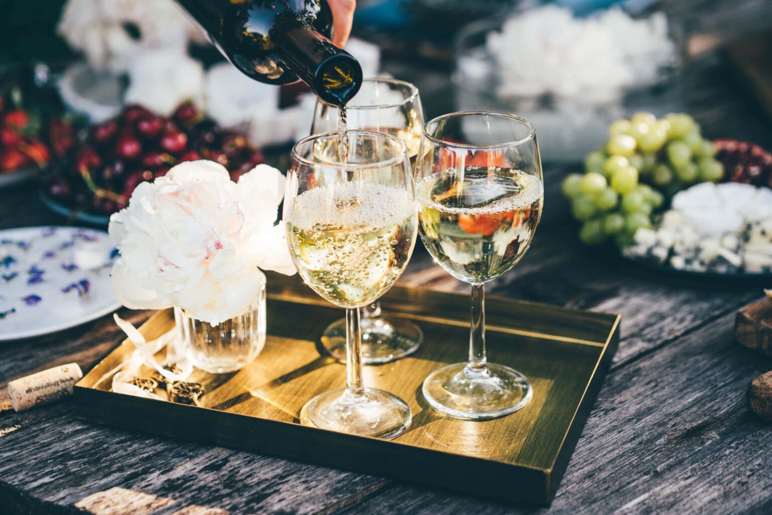 A person pours white wine into three glasses on a brass tray beside a white flower in a vase, with plates of grapes, cheese, and cherries in the background.