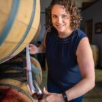 A woman pours wine from a wooden barrel into a glass while smiling and standing in a winery.