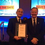 Two individuals stand together indoors, both smiling. One holds a framed certificate labeled "Decanter Scholarship." A screen in the background displays information about a WSET graduation event.