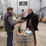 Two men in a wine distillery sample wine from a bottle while standing beside a barrel. Large steel tanks and wooden barrels are visible in the background.