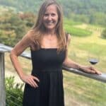 A woman in a black dress stands by a wooden railing holding a wine glass, with a scenic vineyard and hills in the background.