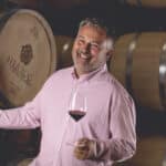 A man in a pink shirt smiles while holding a glass of red wine in a winery. Large wine barrels are visible in the background.