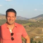 A man in a pink shirt stands smiling in front of a scenic background with hills and terraced vineyards under a clear sky.