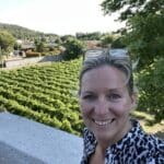 A woman with blond hair smiles for a selfie overlooking a lush vineyard under a clear sky.